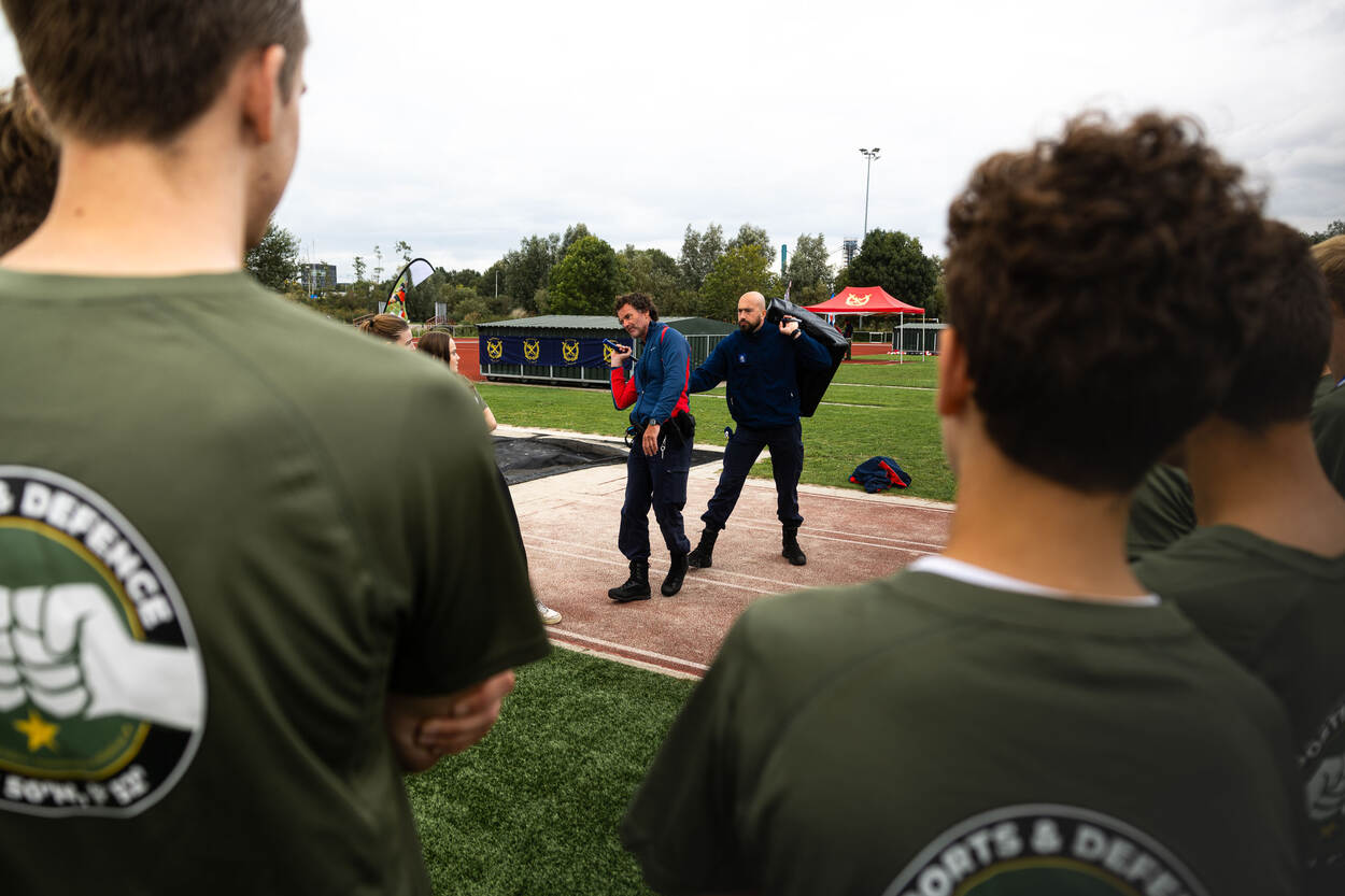 Een man wordt van achteren aangevallen en verdedigt zich. Op de voorgrond aandachtige scholieren.