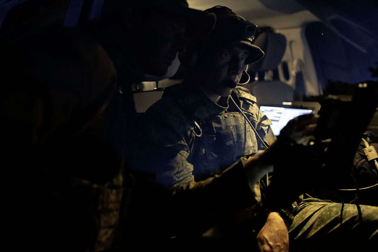 Twee militairen kijken naar een scherm. De ene zit in het voertuig, de ander staat ernaast. De foto is van boven de openstaande autodeur genomen, in de duisternis.