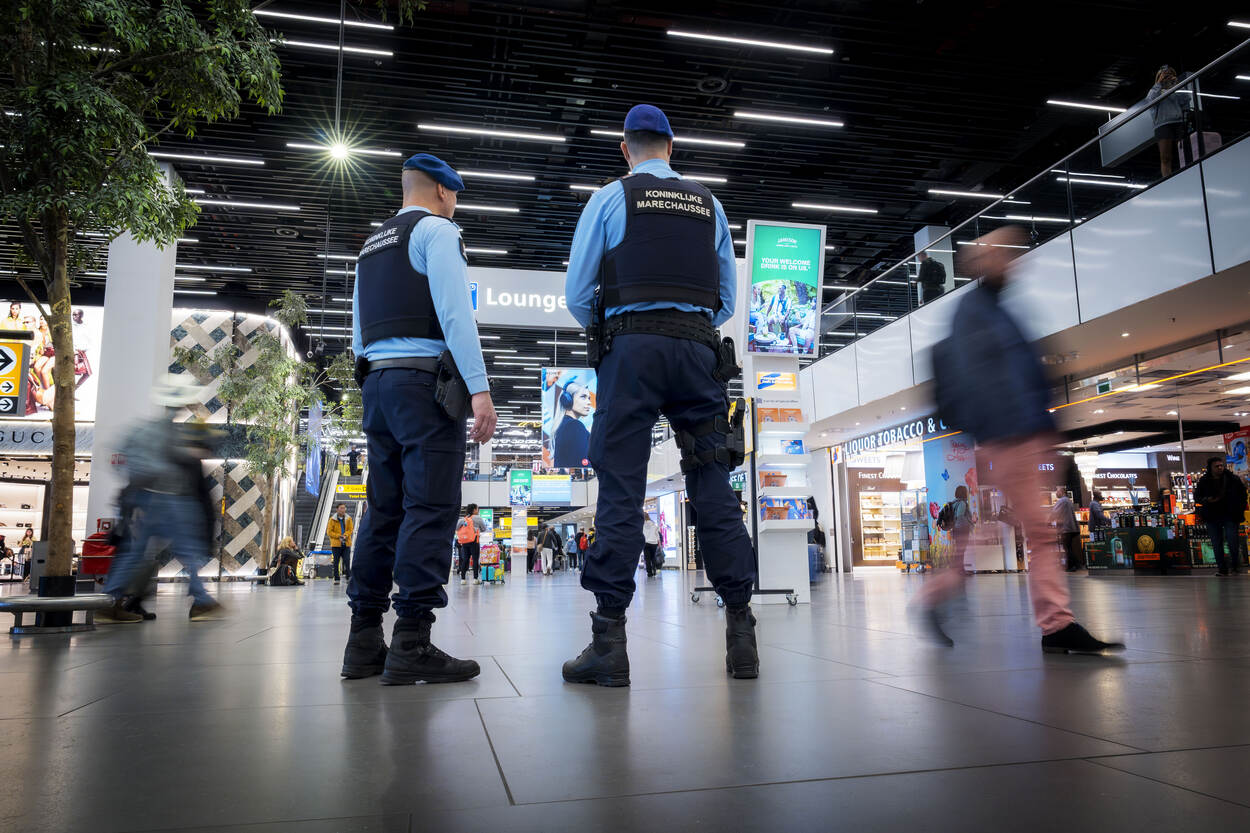 Twee marechaussees staan in de centrale hal van luchthaven Schiphol. Er staat ‘marechaussee’ op hun rug. Om hen heen lopen reizigers.