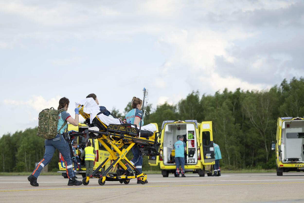 Twee ambulancemedewerkers verplaatsen een jongeman op een rijdende brancard. Een van hen draagt een rugzak van Defensie. Op de achtergrond staan meerdere ambulances opgesteld.