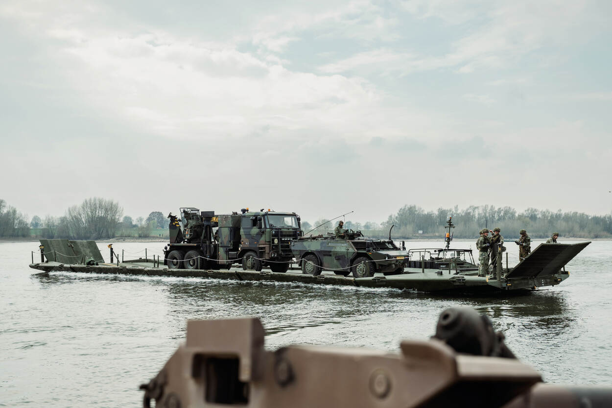 Een drijvende brug vervoert militaire voertuigen over de Duitse Rijn. Op het vaartuig staan militairen die het proces begeleiden.