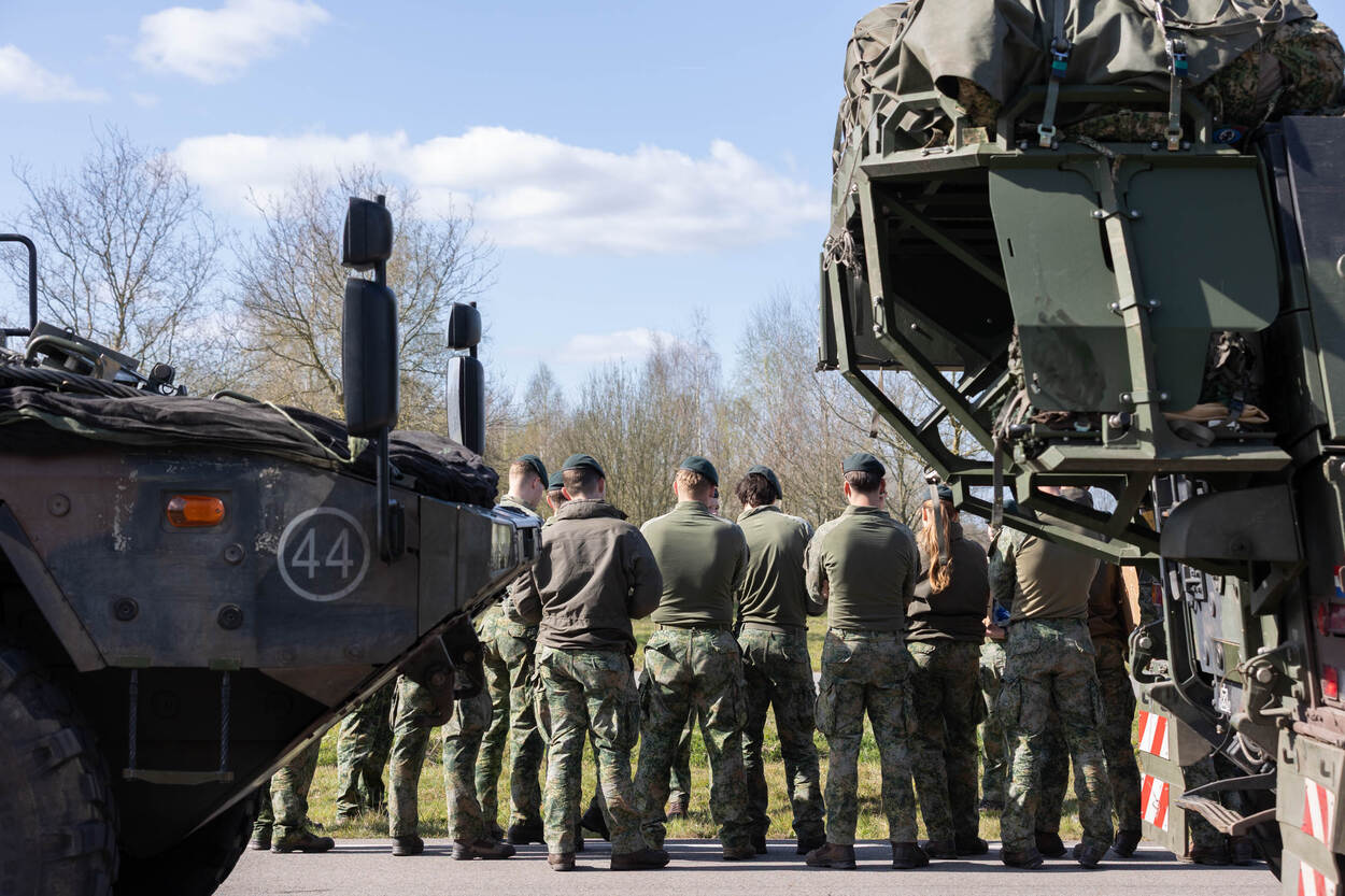 Groep militairen op de rug gezien overleggen naast geparkeerde voertuigen.