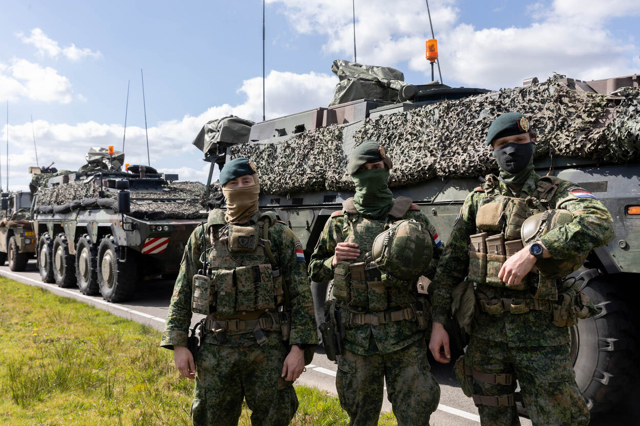 3 militairen met gezichtsbedekking staan voor geparkeerde voertuigen.