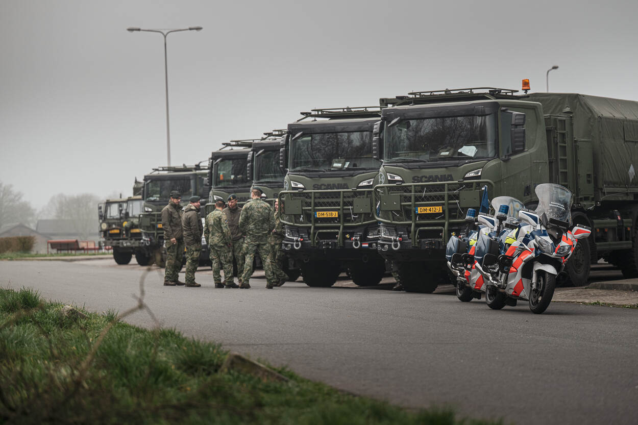 Militaire vrachtwagens naast elkaar geparkeerd. Een groepje militairen staat voor de wagens te praten. Op de voorgrond staan twee motoren van de marechaussee.