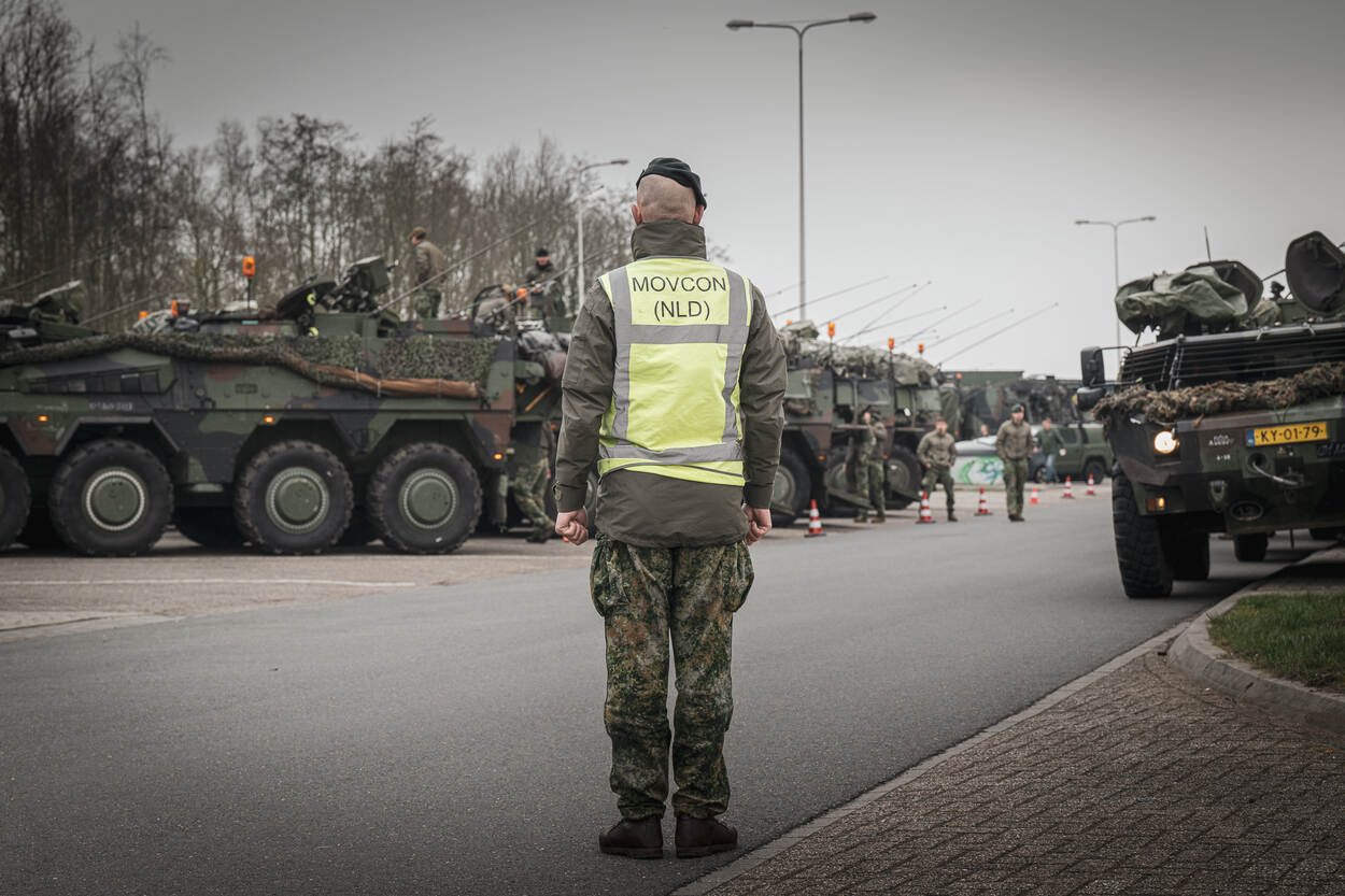 Militair met geel hesje staat op parkeerplaats tussen militairen voertuigen.