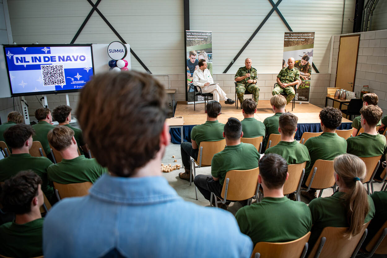 Twee militairen zitten met presentator op podium in zaal vol studenten.
