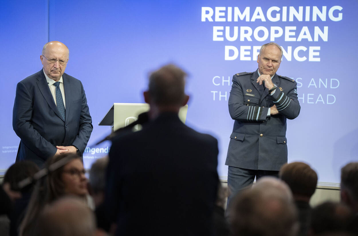 Man in pak en militair staan op podium voor volle zaal.