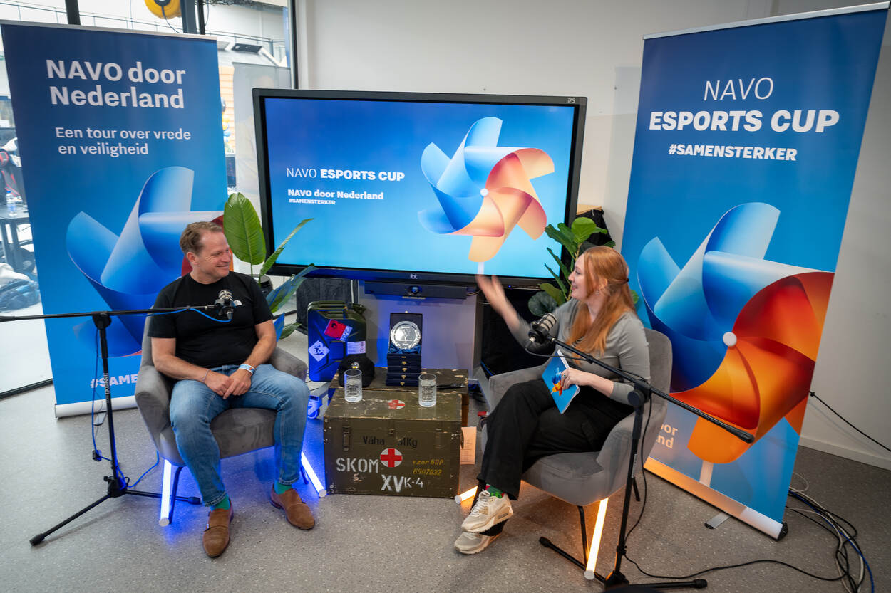 Gast in studio wordt geïnterviewd. Beide personen zitten op stoel voor een beeldscherm en banners.