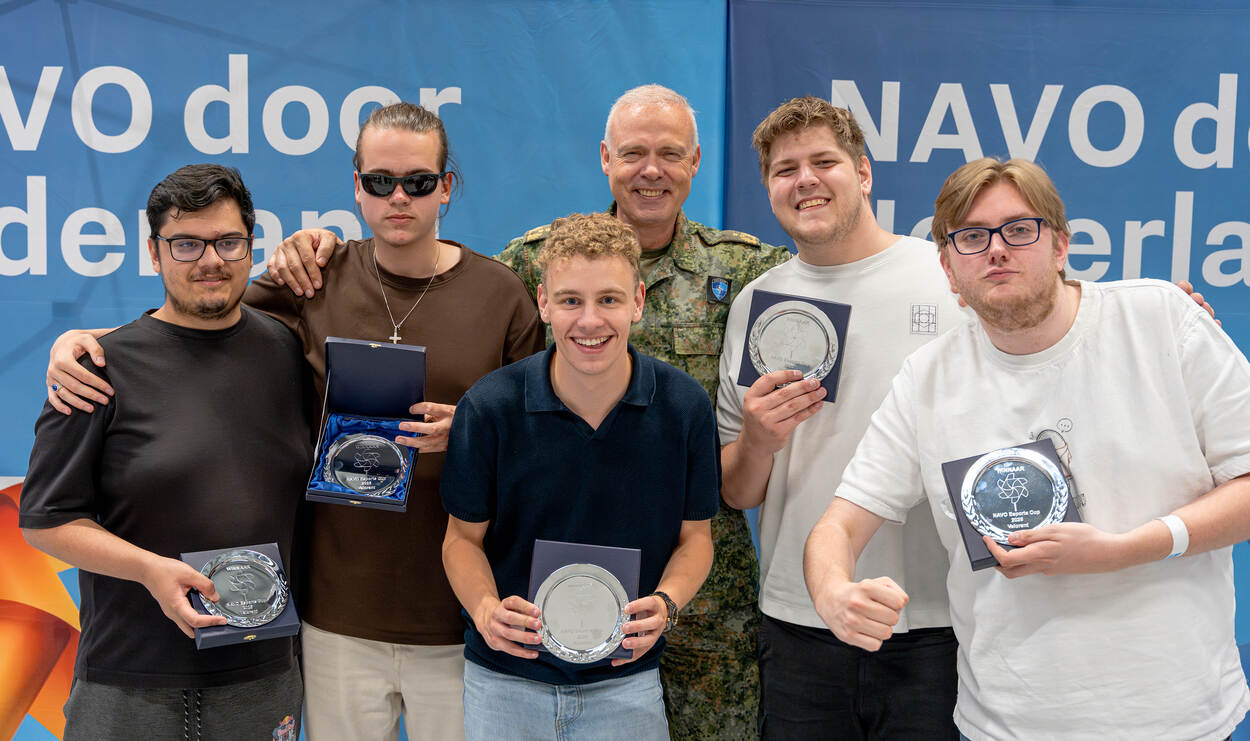 5 jongeren en 1 militair staan lachend naast elkaar. De jongeren houden een kampioenschaal vast.