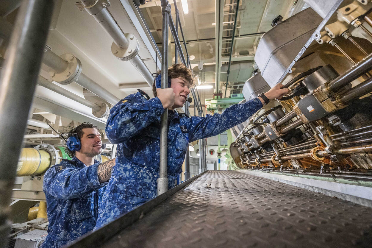 Een militair in een blauw vlekkentenue buigt voorover naar een buizeninstallatie, geflankeerd door een collega.