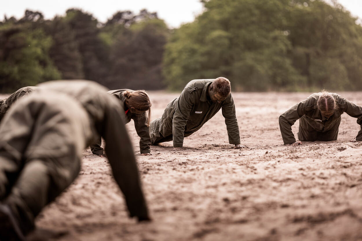 Een groep mensen ligt in de voorligsteun in het mulle zand. Ze dragen groene overalls.