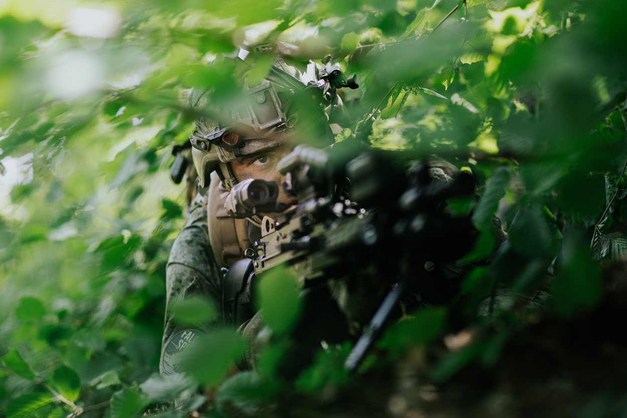 Een gecamoufleerde militair in de bosjes. Hij houdt een lang vuurwapen vast.