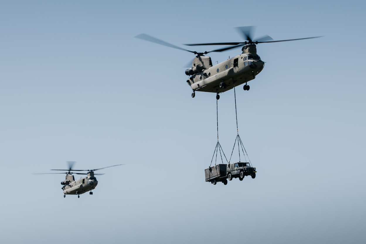 Twee Chinook-helikopters vliegen achter elkaar door de lucht. Een daarvan verplaatst een auto, die onder de helikopter hangt aan een touw.