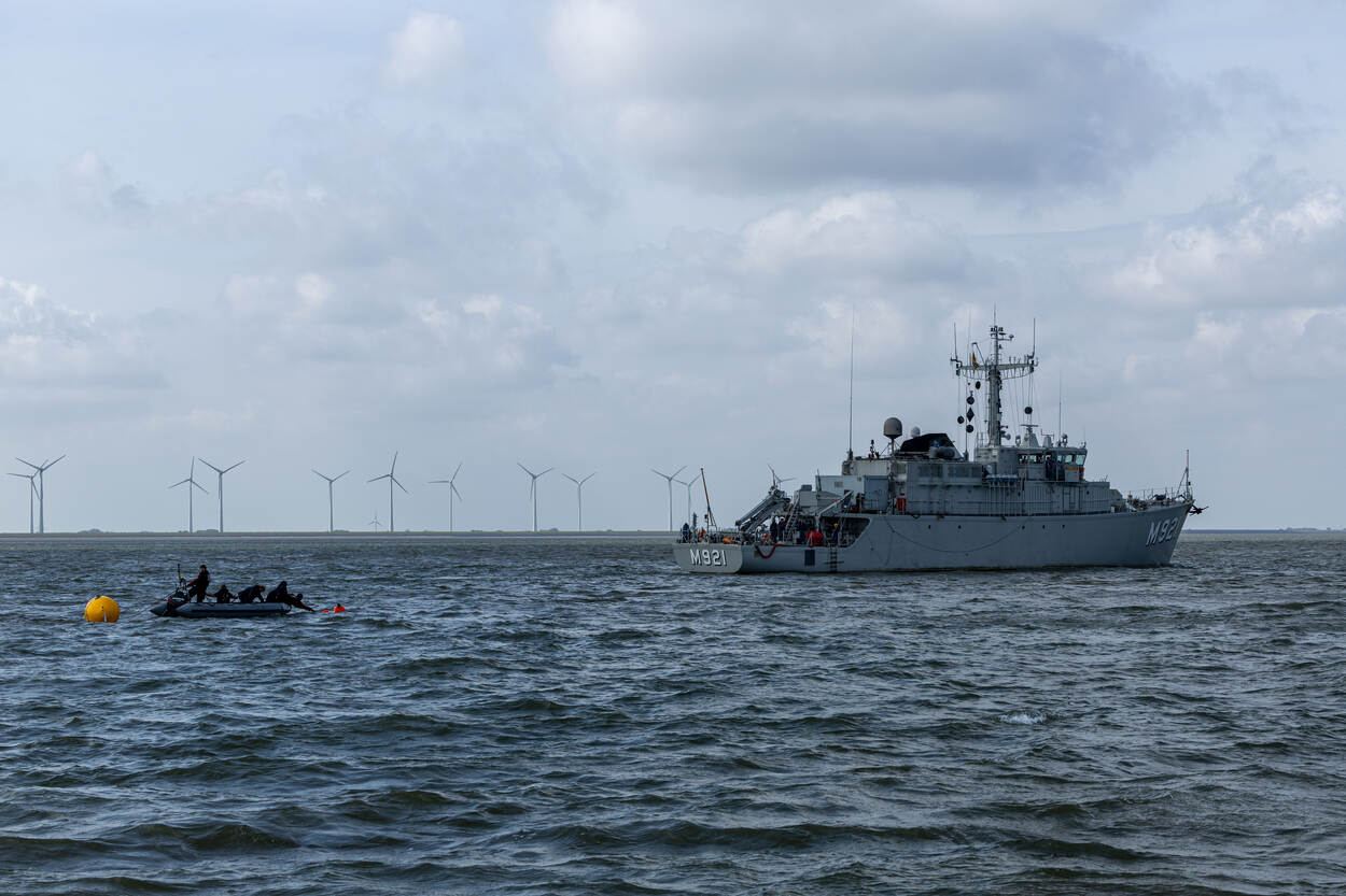 Vanuit een rubber bootje zetten duikers een boei uit, rechts is BNS Lobelia te zien en op de achtergrond de kust van Eemshaven met een aantal windturbines.