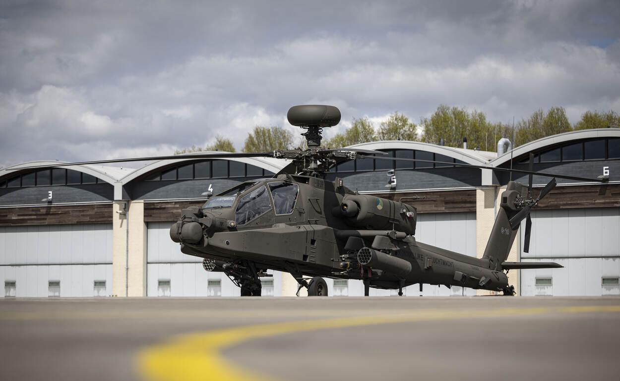 De donkergroene Apache-helikopter staat stil op de grond. Daarachter zijn de hangaars te zien.