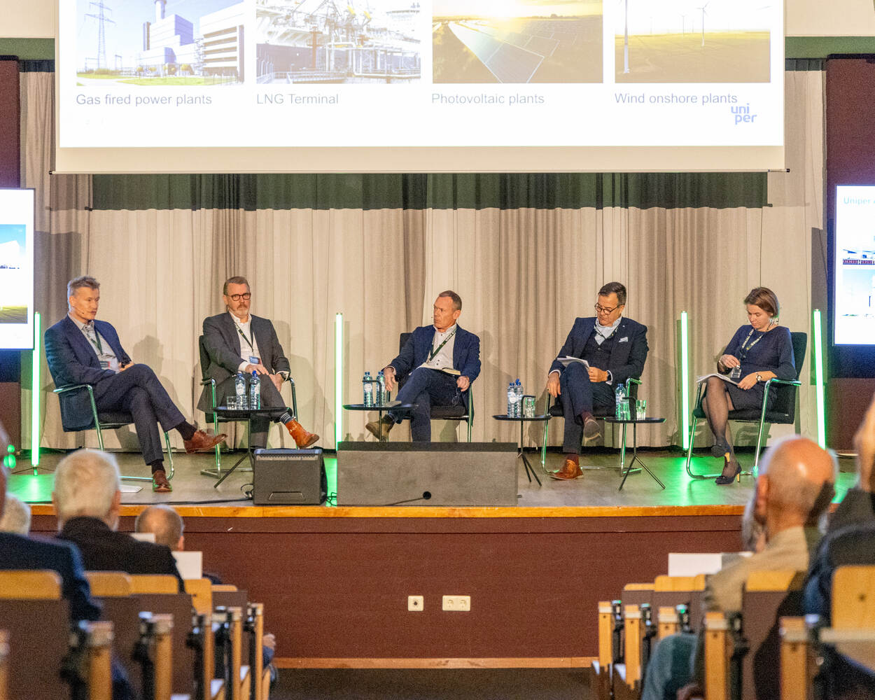 5 personen praten met elkaar op een podium met gordijnen en videobeelden op achtergrond.