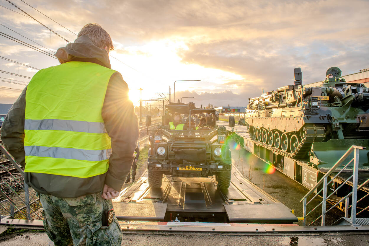 Militair materieel wordt van een trein gereden, op de achtergrond een felle zon.