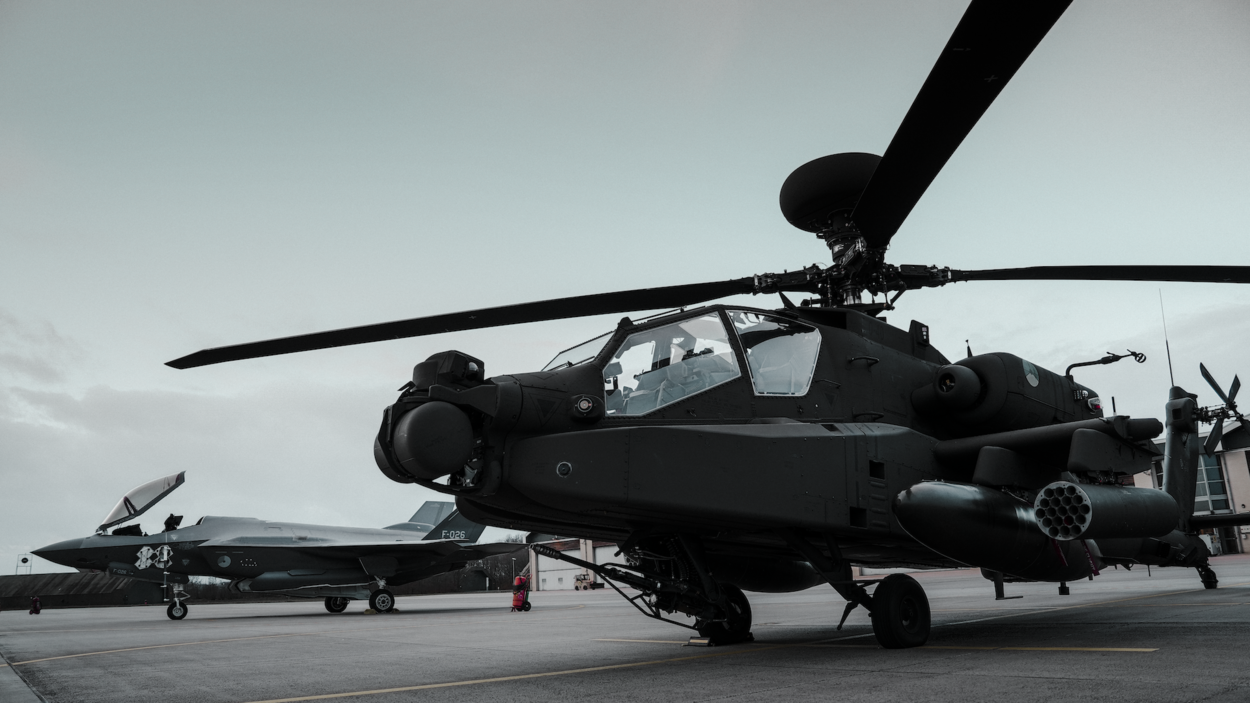 Apache met draaiende rotorbladen staat op een militaire landingsbaan; op de achtergrond is een F-35 geparkeerd onder een bewolkte lucht.