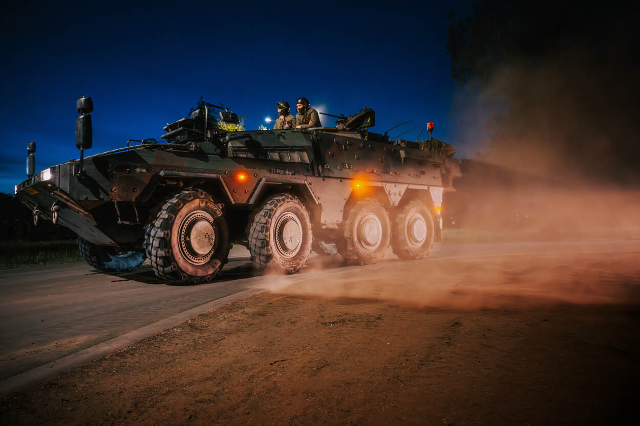 Een Boxer rijdt de weg op en creeert een stofwolk. Bovenluiks kijken militairen mee. Alles gebeurt in de duisternis.