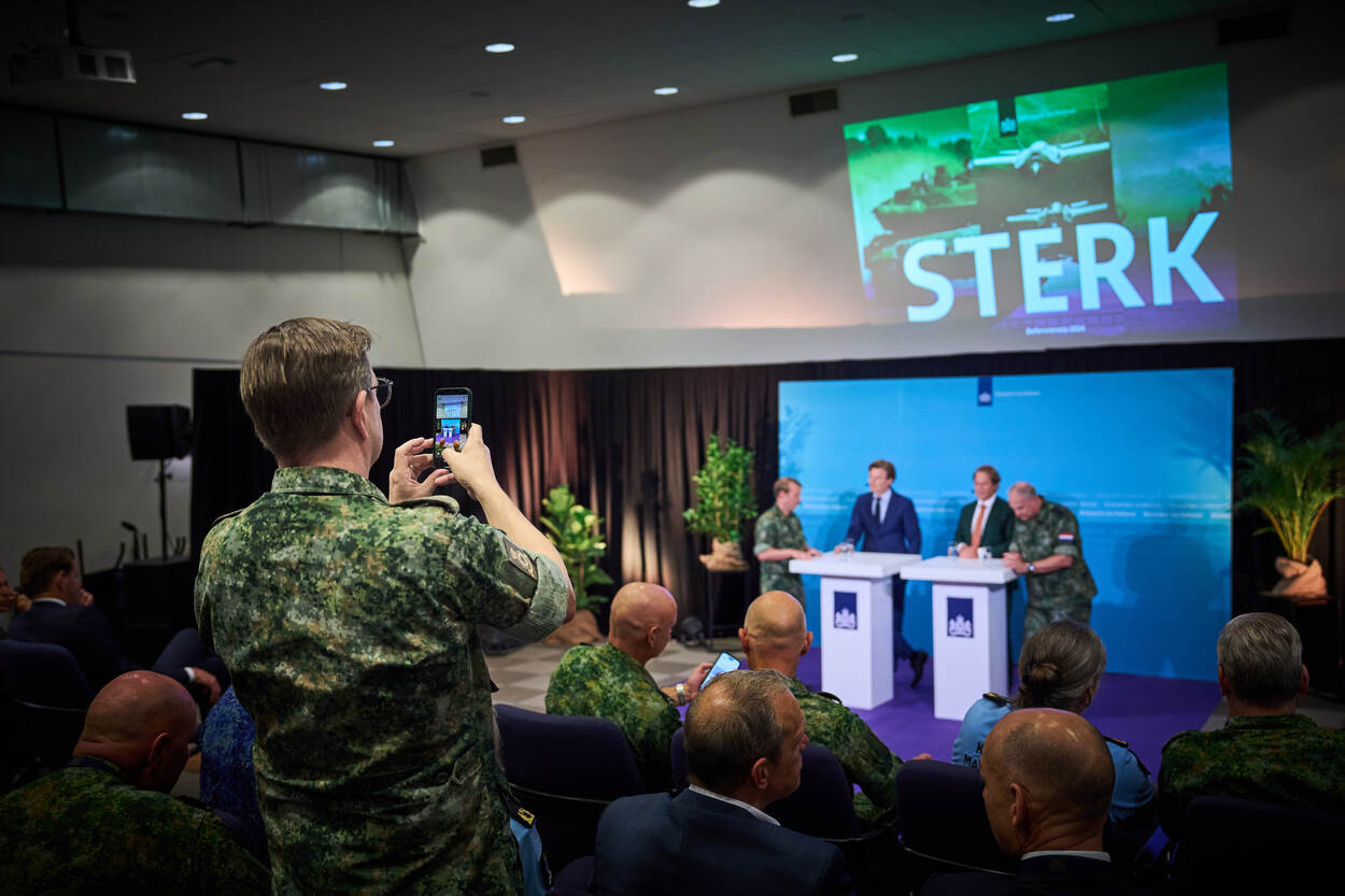 Een militair maakt met zijn telefoon een foto, terwijl de bewindslieden van achter een spreekgestoelte de Defensienota van 2024 presenteren.