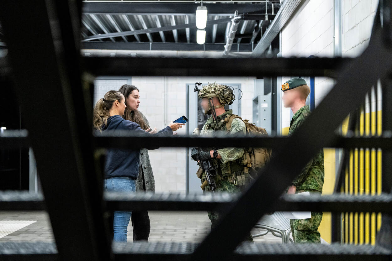 Twee dames staan op een metroperron en interviewen een militair met een telefoon voor hun gezicht.