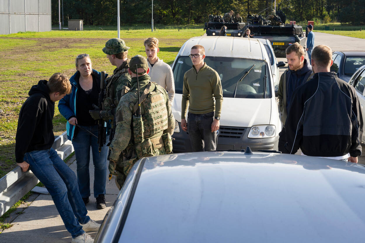 Een jongen hangt op de vangrail, omgeven door andere burgers, lopend in de file. Daarachter staan twee Fennek-verkenningsvoertuigen geparkeerd.