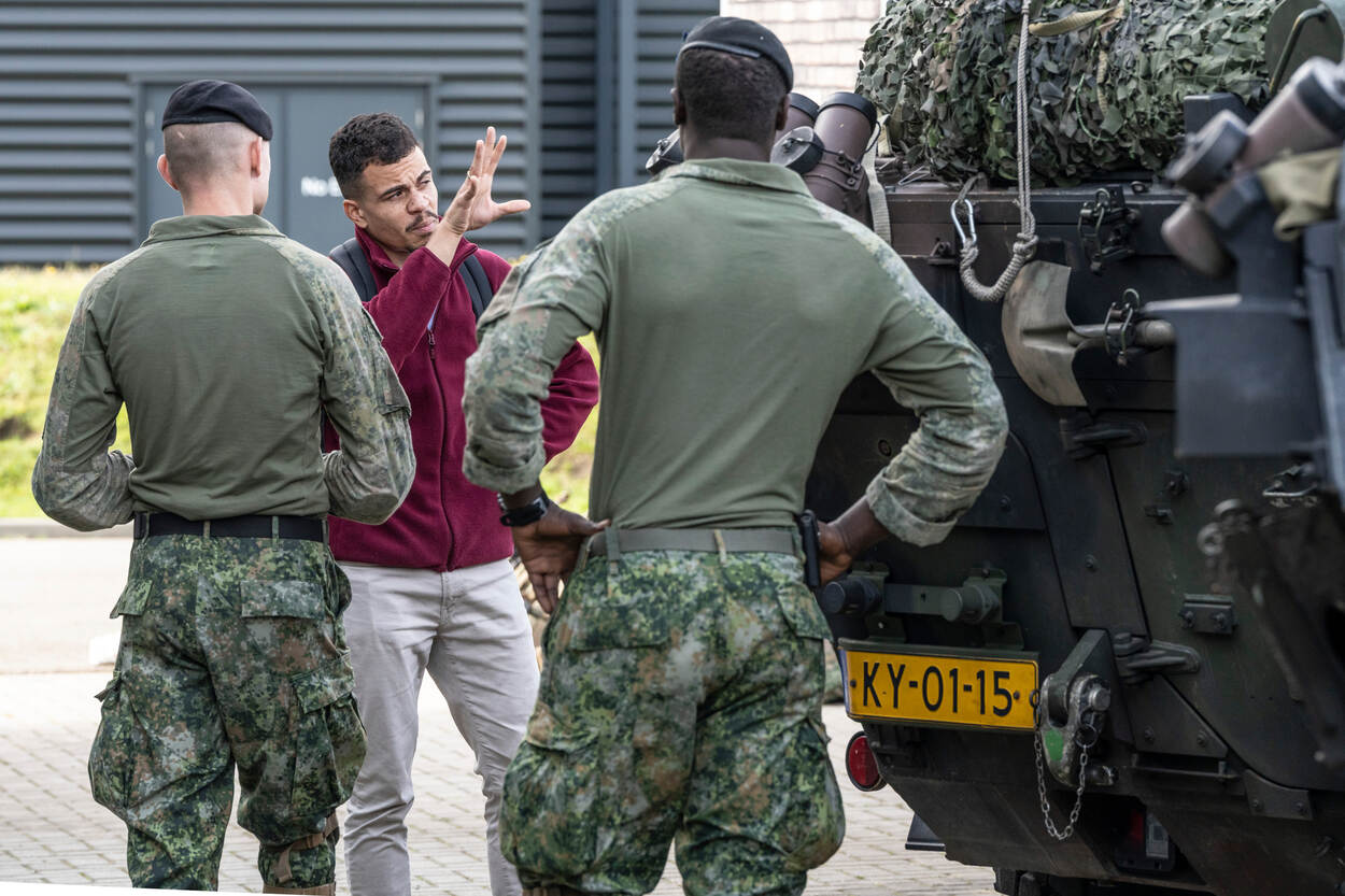 Twee militairen praten met een acteur in een fictieve winkelstraat tijdens een statische show, staand naast hun verkenningsvoertuig, de Fennek.