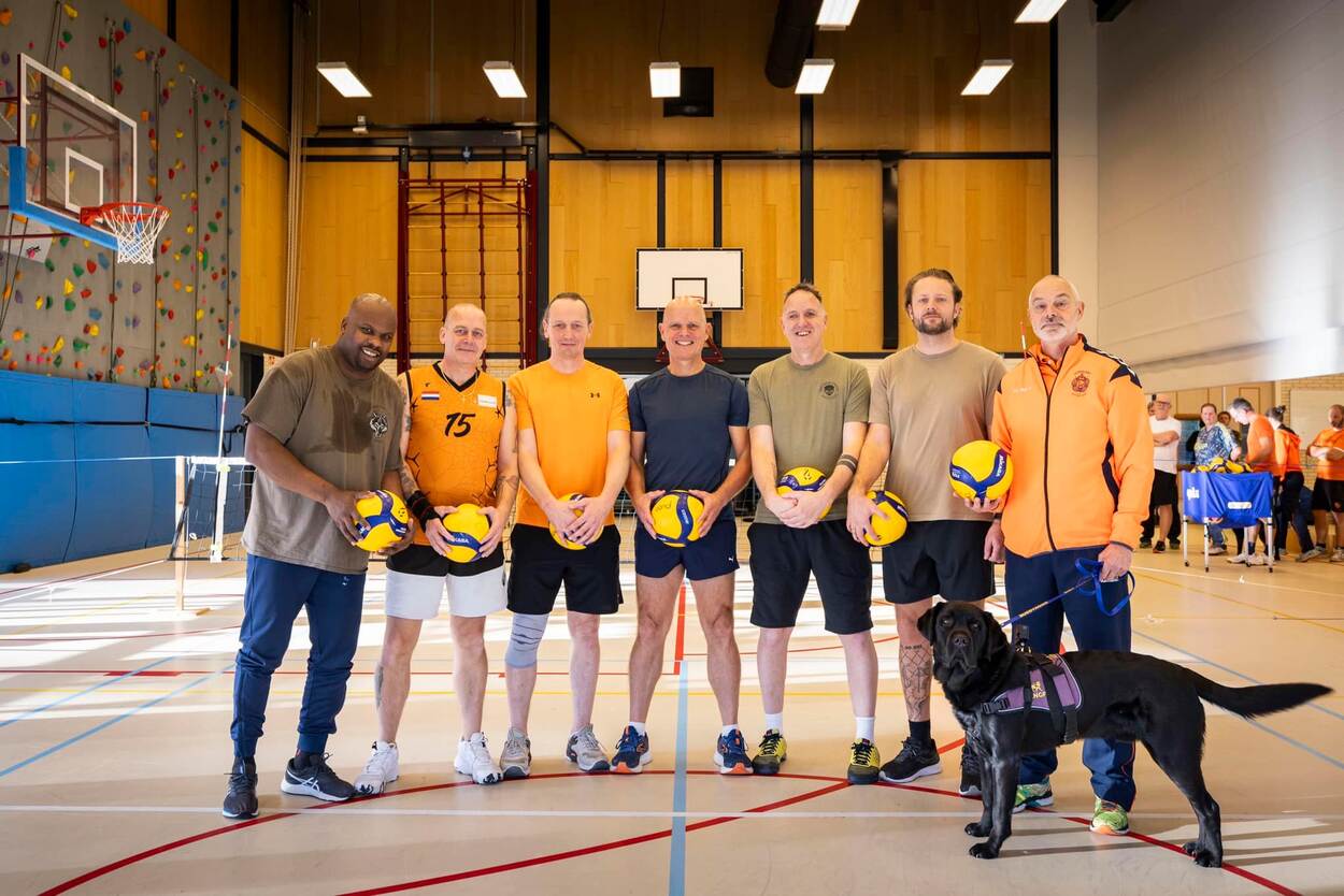 Zeven mannen in sportkleding op een rij in een sportzaal met een bal in hun handen. De man rechts wordt vergezeld door een zwarte hulphond.