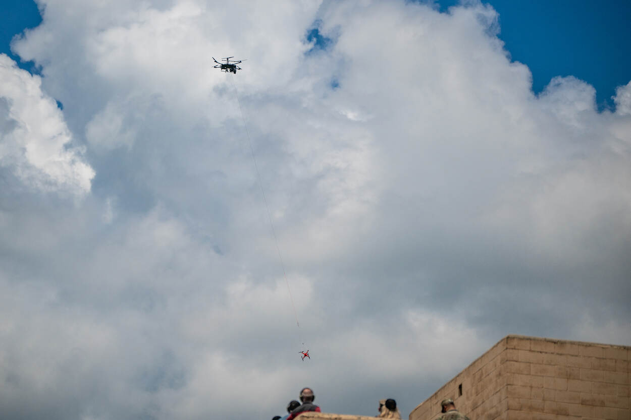 Foto links: een viertal militairen kijkt in de camera. Voor hen staan witte, grote drones. Foto rechts: een drone vliegt door de lucht, op het dak van een gebouw kijken mensen naar het vliegende vaartuig.