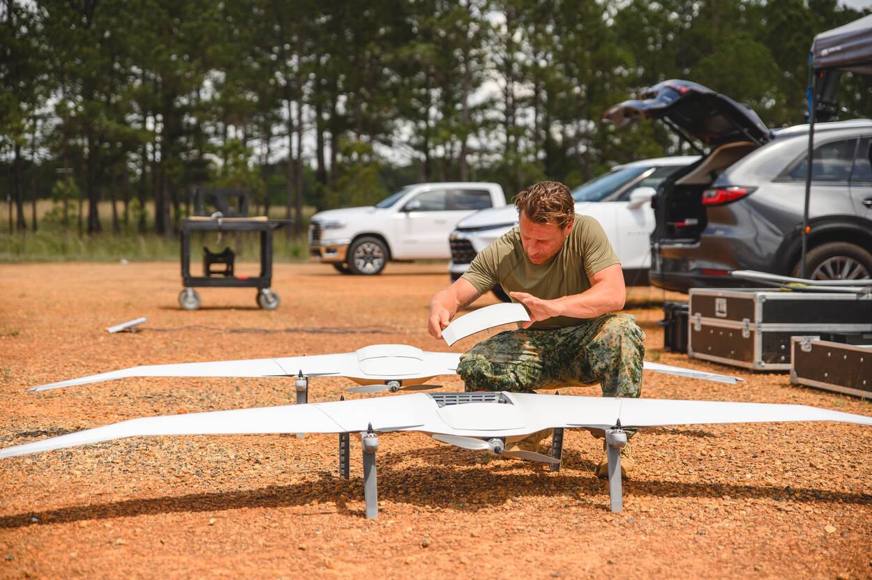 Foto links: korporaal Apeldoorn sleutelt aan een grote witte drones. Foto rechts: een groep mensen en militairen onder witte partytenten, met daaromheen zwarte kisten met grijze randen.