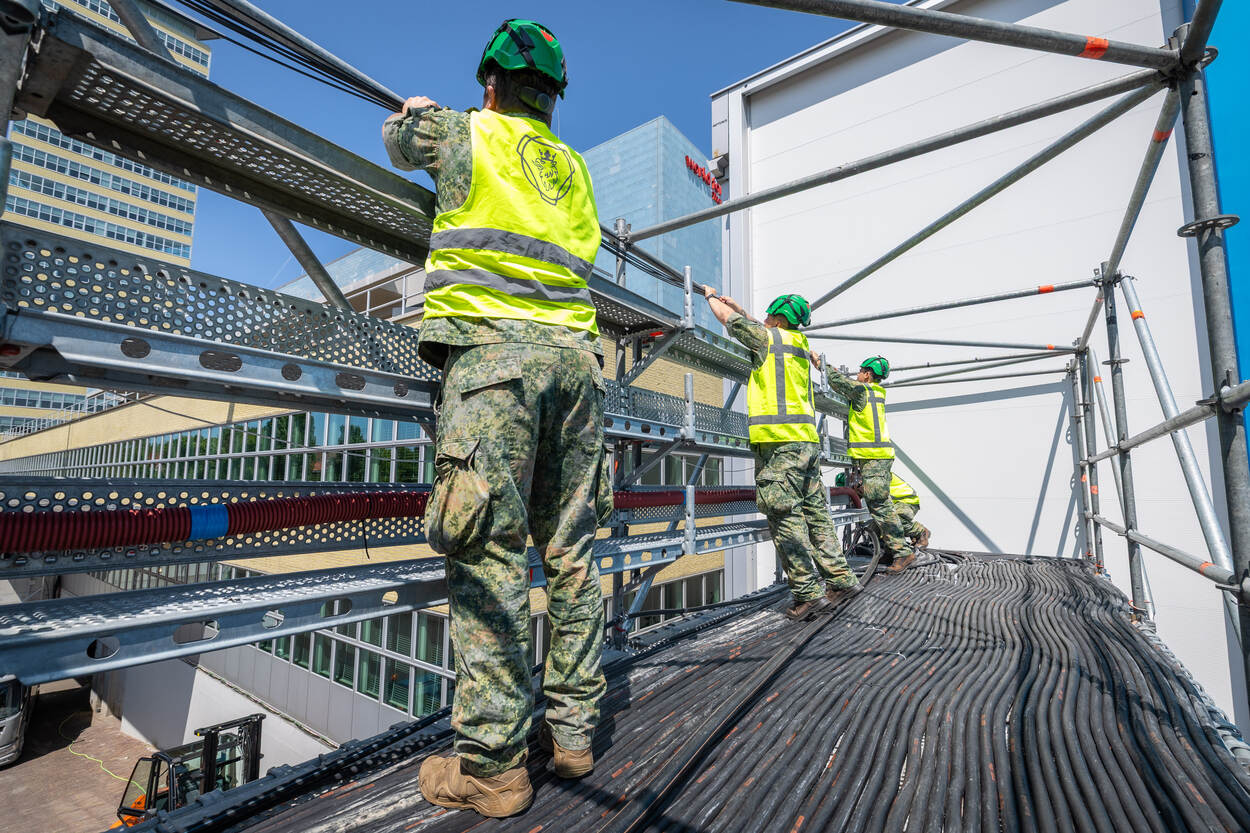 3 militairen in hesjes en met helmen leggen kabels aan op stellage.