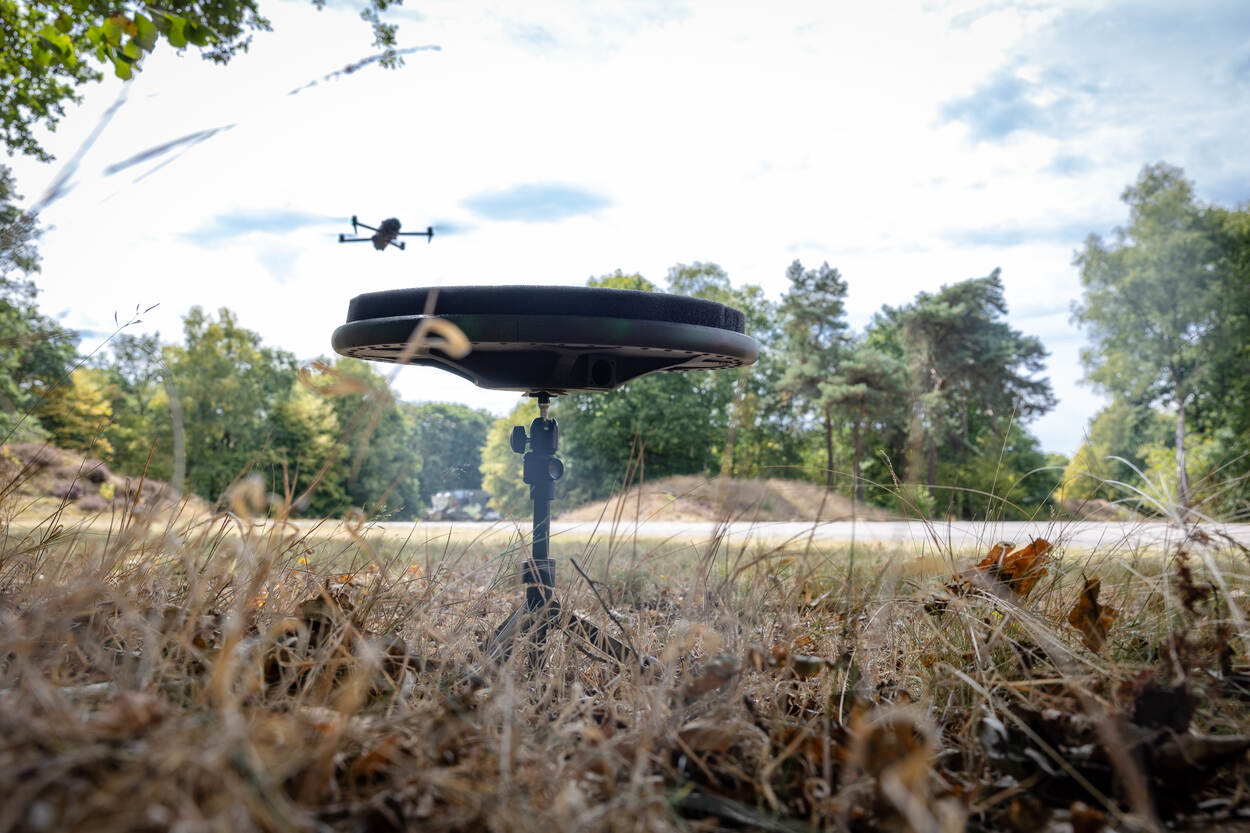Een sensor op een driepoot opgesteld in het groen. Erboven hangt een drone.