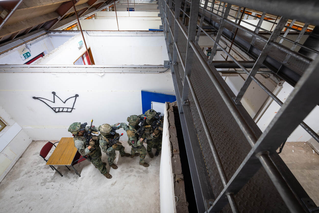 Vier militairen van bovenaf gezien die tijdens een oefening vanuit een kamer een gang inlopen.