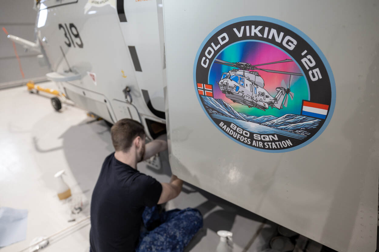 De badge van Cold Viking groot in beeld. Op de achtergrond werkt een technicus aan een NH90.