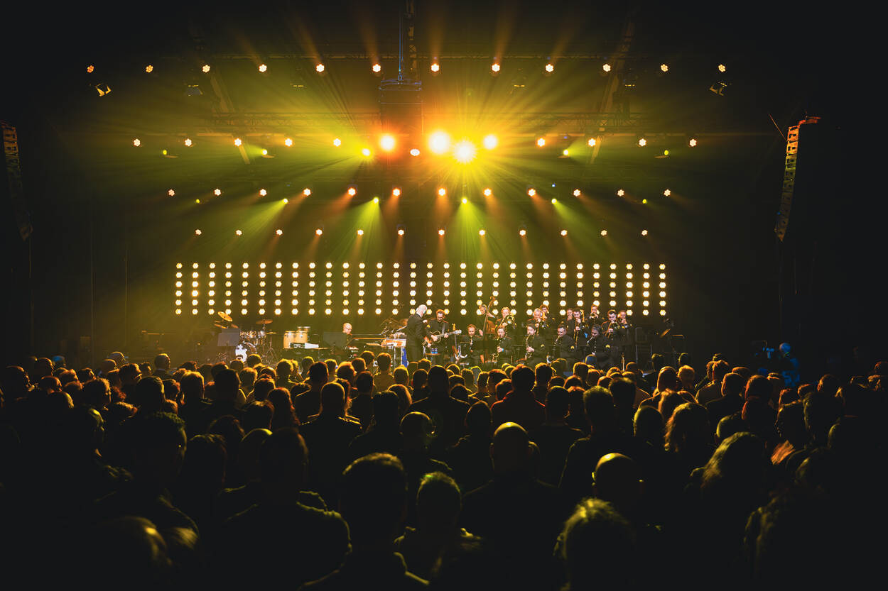 Het Orkest Koninklijke Luchtmacht met daarboven felle lampen en op de voorgrond publiek.