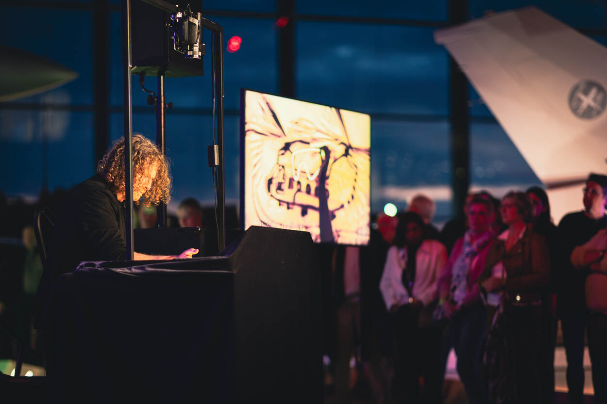 Zandtovenaar Gert van der Vijver aan het werk. Op de achtergrond een tv-scherm waarop te zien is wat hij met zand maakt.