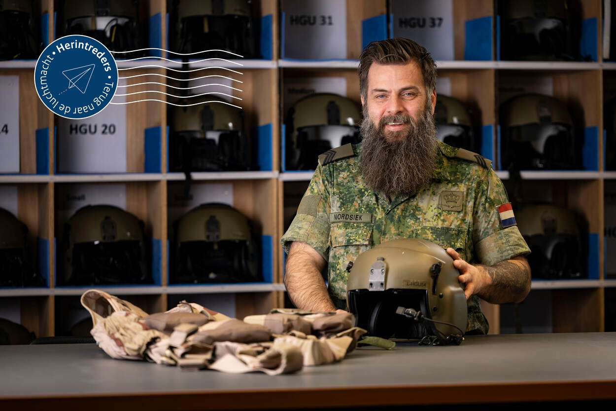 Sergeant Einar Nordsiek poseert achter een tafel met daarop een vliegerhelm en -vest. Achter hem een vakkenkast met vliegerhelmen.