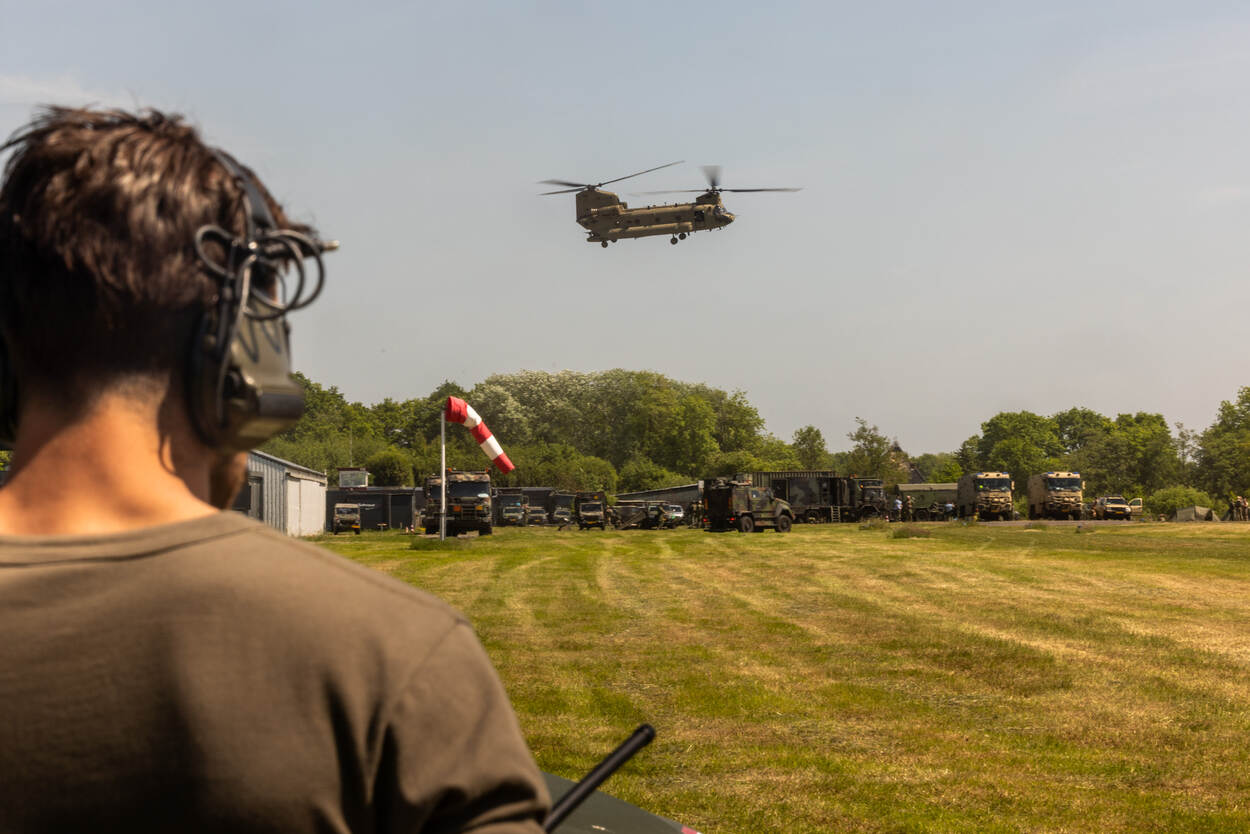 Vliegende Chinook boven geparkeerde militaire voertuigen op een grasveld.