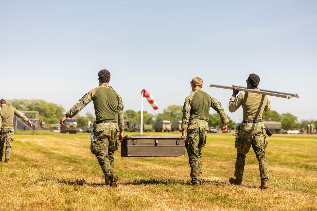 Drie militairen sjouwen met materieel over het gras.