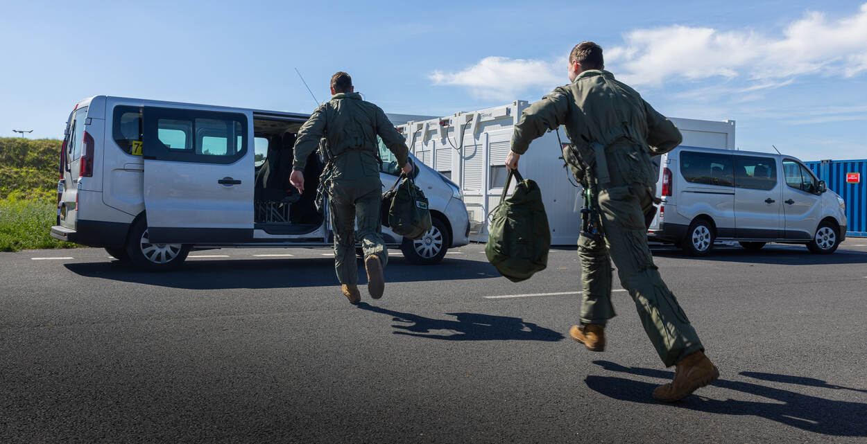 Twee vliegers rennen in flightsuit met helmtas in de hand naar een busje.
