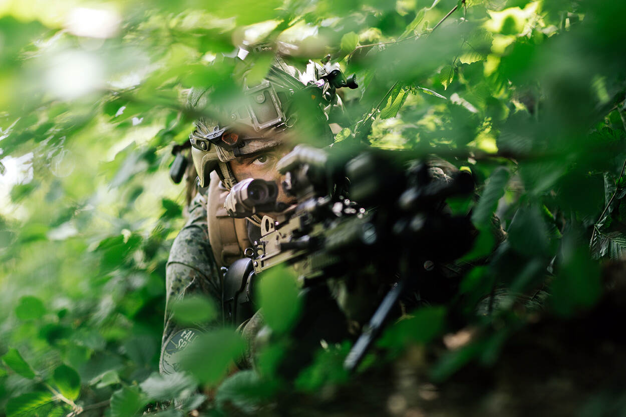Een gecamoufleerde militair in de bosjes. Hij houdt een lang vuurwapen vast.