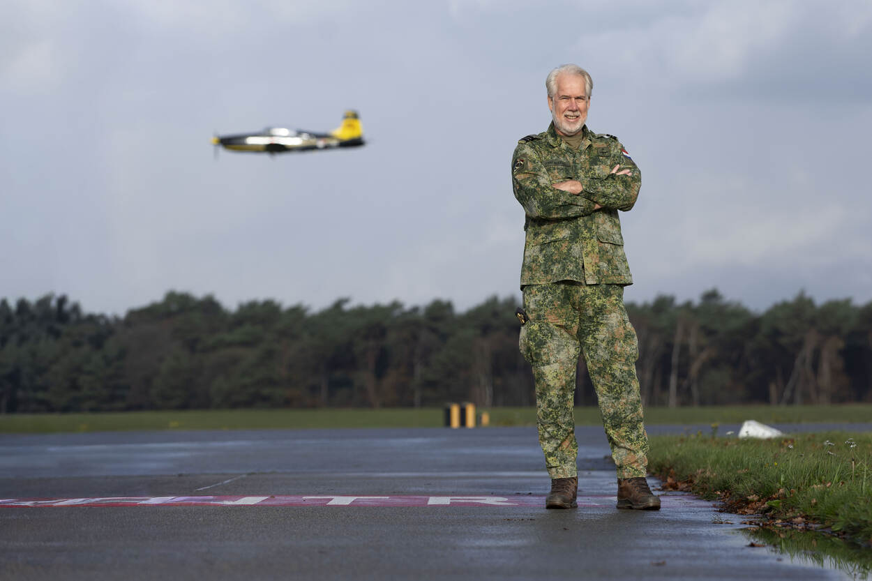 ‘Een militair in camouflagepak staat met armen over elkaar aan de rand van de taxibaan. Op de achtergrond vliegt een PC-7 laag voorbij.