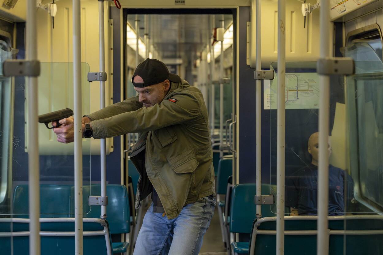 Man met petje op houdt met pistool in beide handen zienderogen iemand onder schot in de metro, op de achtergrond is een passagier zichtbaar.