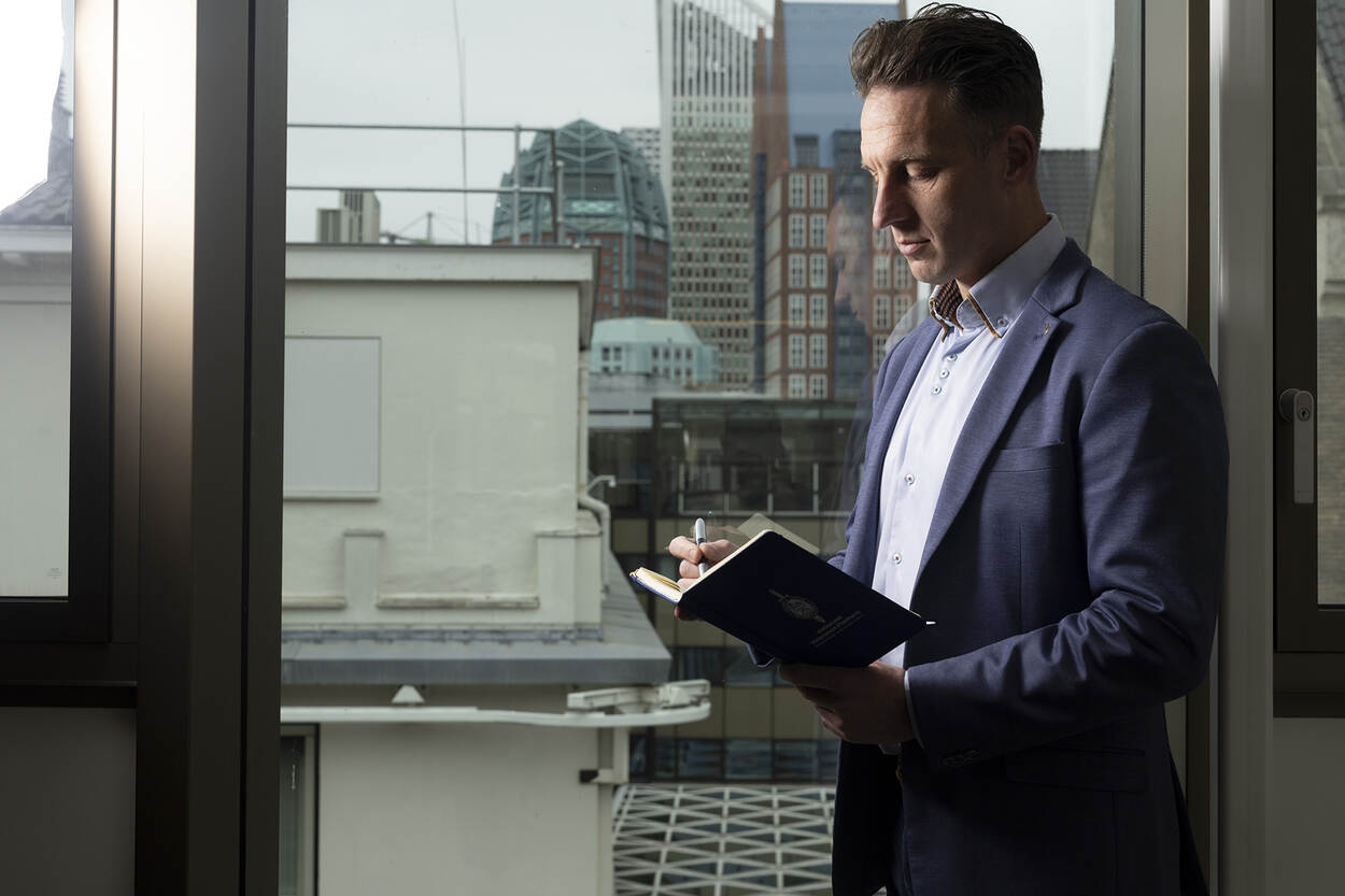 Wietse staat voor een raam en schrijft in een boekje. Op de achtergrond de skyline van Den Haag.