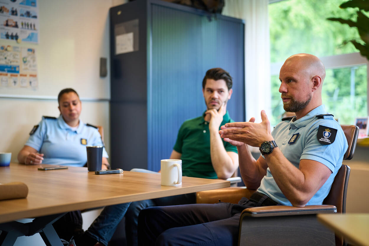 Een kapitein van de KMar geeft uitleg met opengevouwen handen. Zijn collega en een burger luisteren.