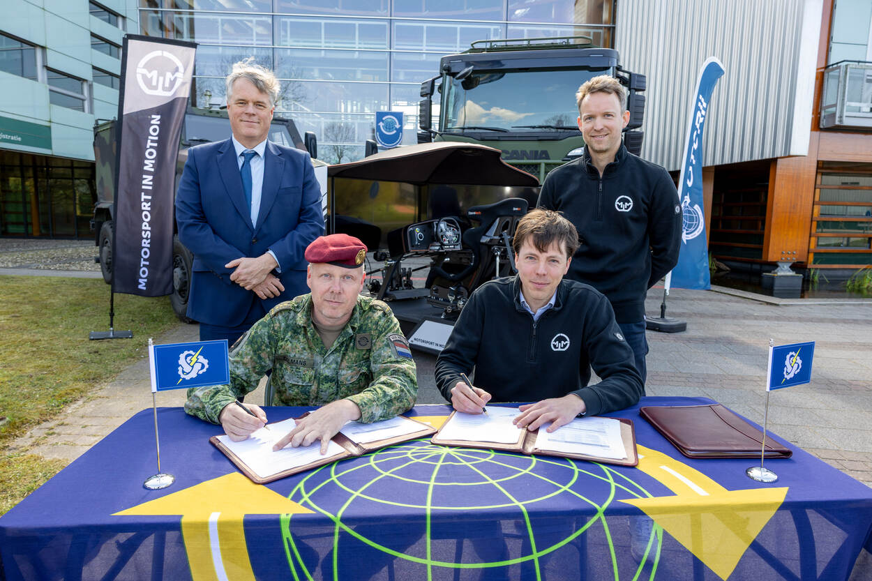 2 mensen tekenen een overeenkomst met op de achtergrond 2 anderen en 2 legervoertuigen.