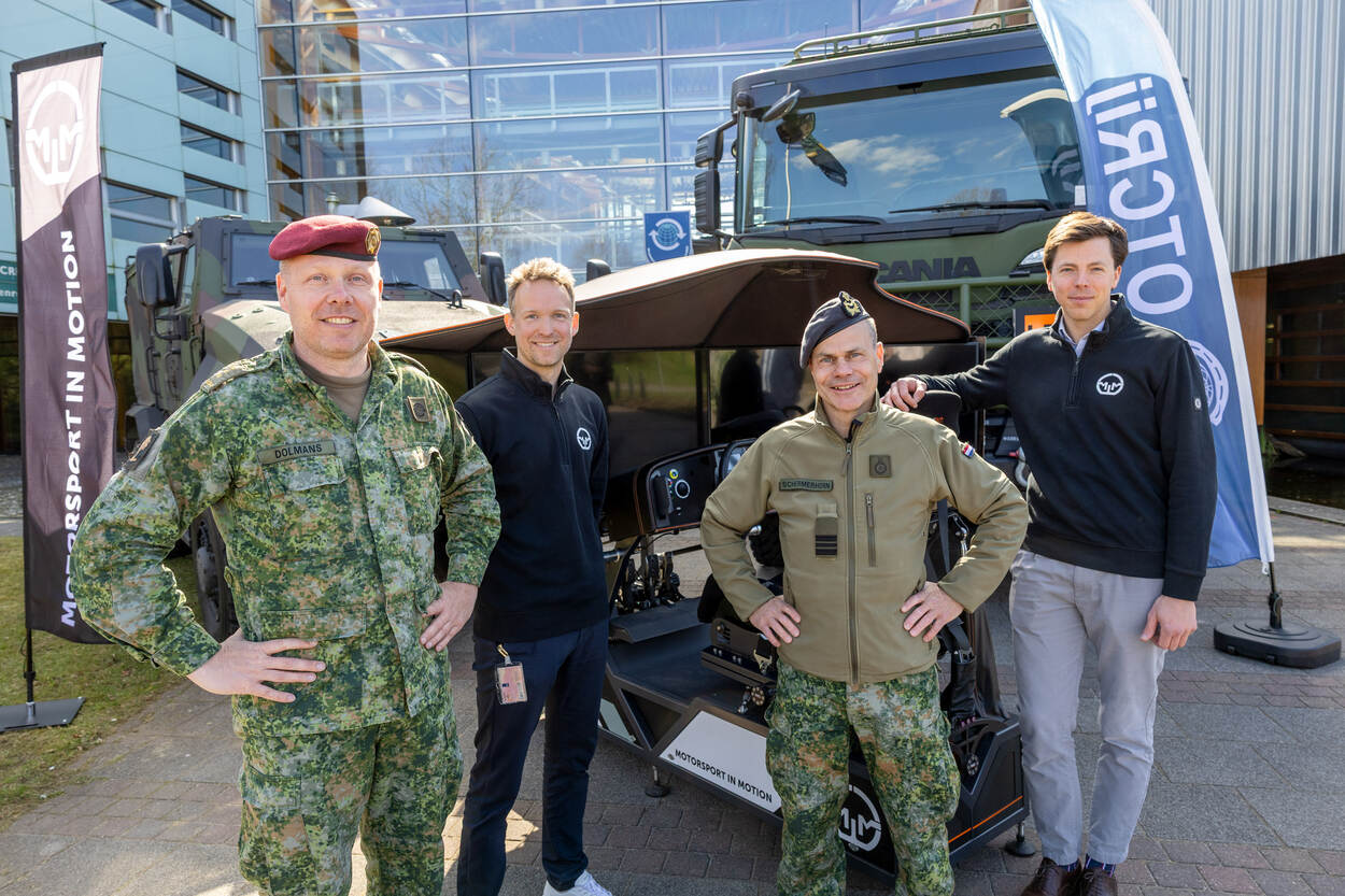 4 mannen poseren voor een simulator en 2 legervoertuigen.