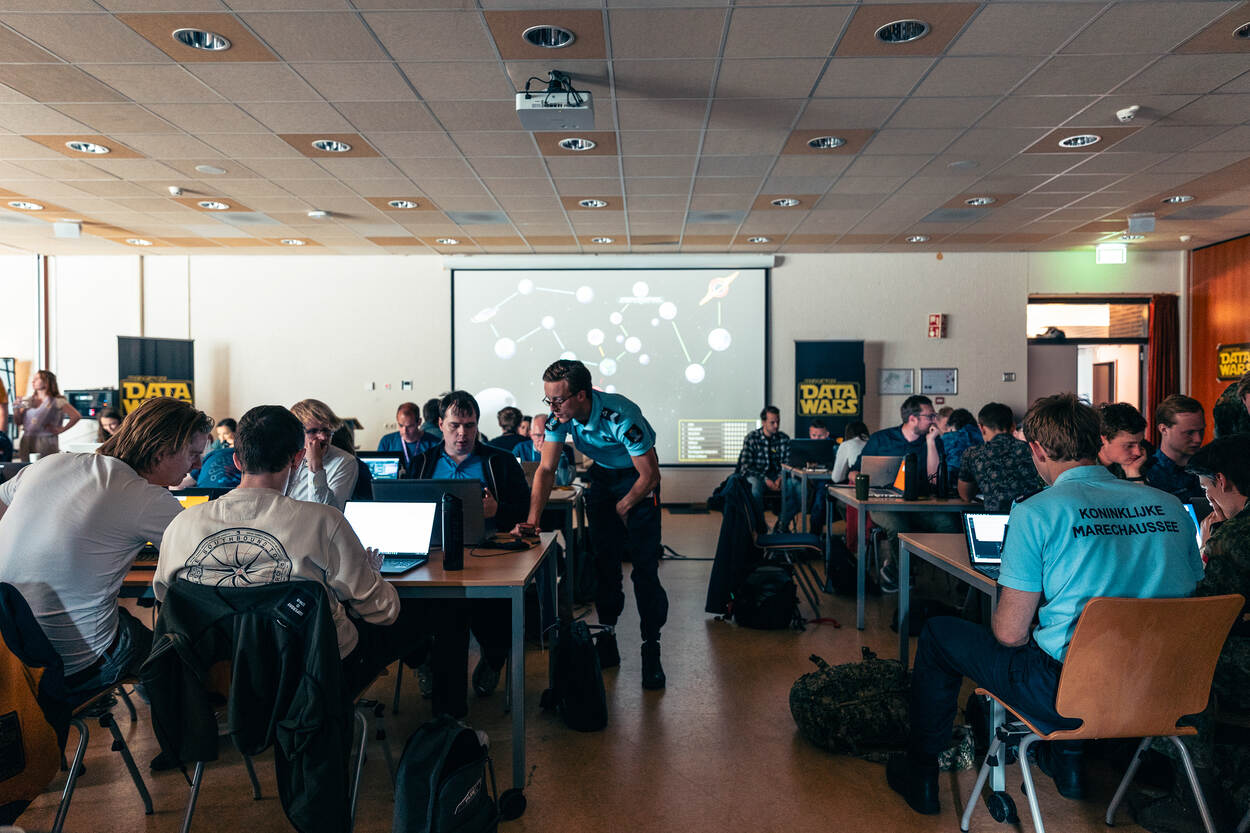 Veel personen zitten in een lokaal achter een laptop met een staand lid KMar en achter een groot videoscherm.