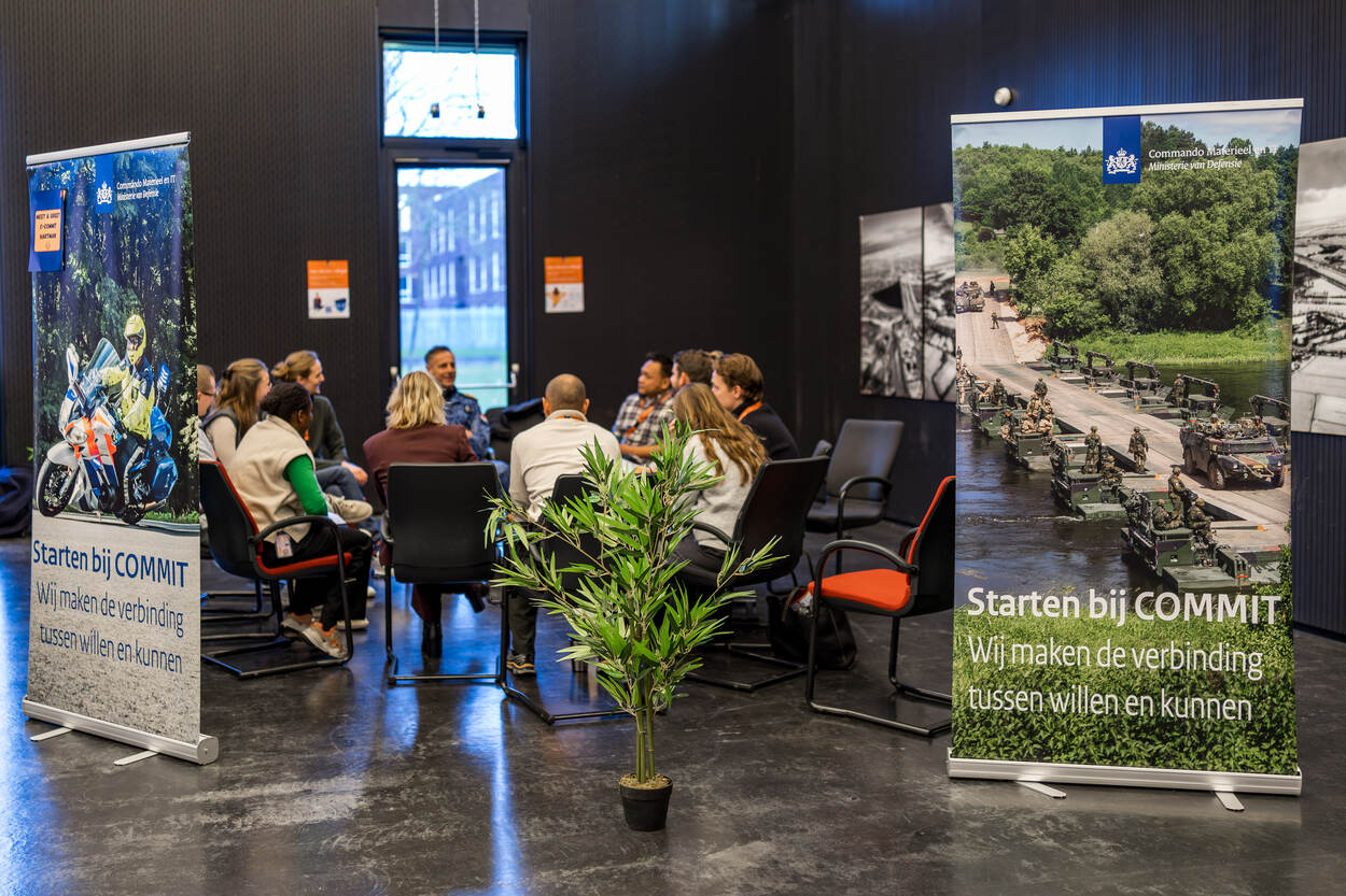 Een groep mensen in een zaal tijdens een presentatie. Twee roll-up banners met tekst en afbeeldingen van COMMIT en Defensie staan prominent weergegeven. Stoelen, planten, en raam op het podium.