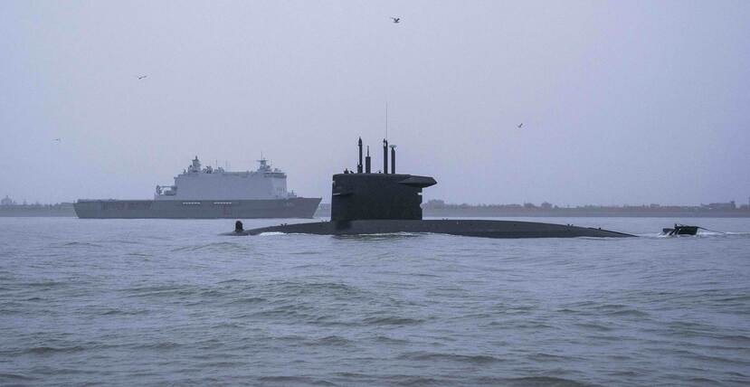 Onderzeeboot half boven water met op de achtergrond een marineschip.